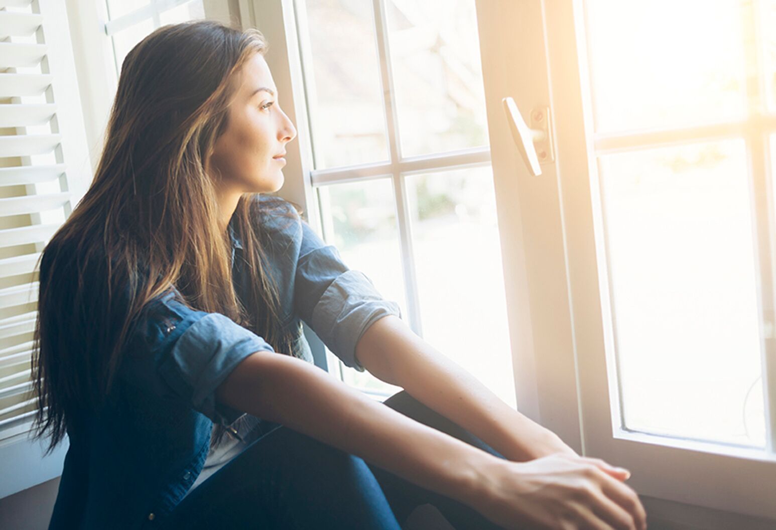 Smiling young woman sitting and looking through the window. With long hair, wears denim shirt. Hands on legs.